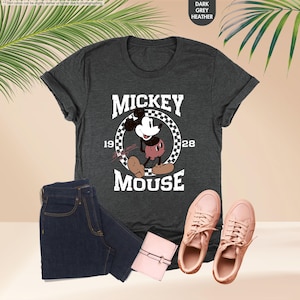 May include: Dark grey heather t-shirt featuring a vintage Mickey Mouse graphic. The design includes the text "MICKEY MOUSE 1928" in a circular frame. The image also shows dark blue jeans, a pink wallet, and pink sneakers.