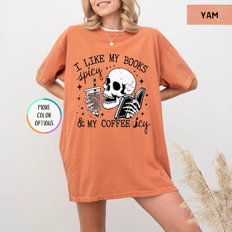 May include: A yam-coloured t-shirt featuring the text "I like my books spicy & my coffee icy" and a skull graphic holding a book and a drink. The shirt has short sleeves and a loose fit.