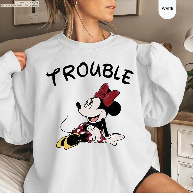 May include: White sweatshirt featuring the word "TROUBLE" in black script above a cartoon image of Minnie Mouse. Minnie Mouse is wearing a red bow, a red and white polka dot dress, and yellow shoes. The sweatshirt has long sleeves.