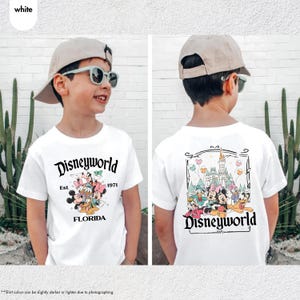 May include: White t-shirt featuring a Disneyworld design. The front of the shirt displays the text "Disneyworld Est 1971 FLORIDA" with cartoon characters. The back of the shirt has a Disney castle and characters. The shirt is worn by a person.