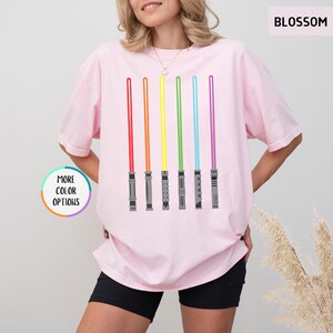 K&ouml;nnte beinhalten: Hellrosa T-Shirt mit einem Aufdruck von sechs farbigen Lichtschwertern in Rot, Orange, Gelb, Gr&uuml;n, Blau und Lila. Das Shirt hat den Text "BLOSSOM" oben rechts und "MORE COLOR OPTIONS" links.