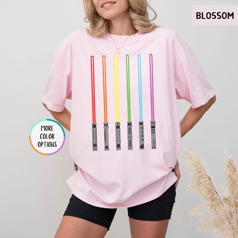 K&ouml;nnte beinhalten: Hellrosa T-Shirt mit einem Aufdruck von sechs farbigen Lichtschwertern in Rot, Orange, Gelb, Gr&uuml;n, Blau und Lila. Das Shirt hat den Text "BLOSSOM" oben rechts und "MORE COLOR OPTIONS" links.