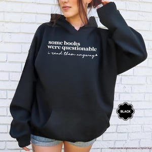 May include: A black hooded sweatshirt featuring white text on the chest that reads some books were questionable i read them anyway. The garment includes a front kangaroo pocket, long sleeves with ribbed cuffs, and a drawstring hood for a casual look.