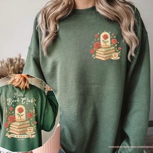 Puede incluir: Sudadera verde militar con un gr&aacute;fico de una rosa en una c&uacute;pula de cristal sobre libros, rodeada de rosas rojas y una taza de t&eacute;. La parte trasera de la sudadera muestra el texto "Belle's Book Club" y "Tale as Old as Time."