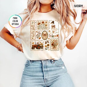 May include: An ivory short-sleeve t-shirt featuring a graphic print with the text hello fall. The design includes illustrations of a tractor, a duck wearing a hat, sunflowers, pumpkins, autumn leaves, and a bow, arranged in rectangular frames.