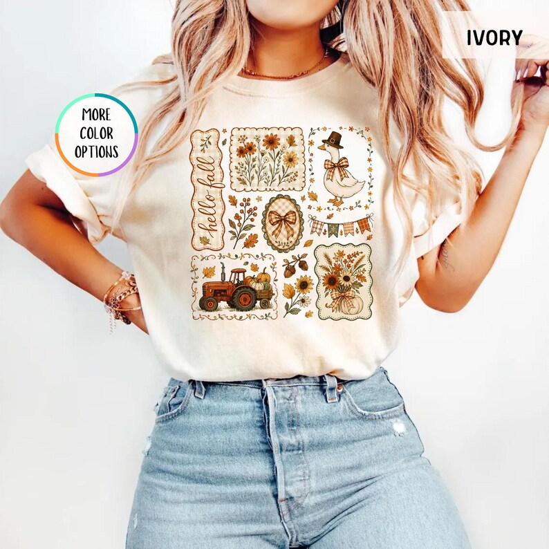 May include: An ivory short-sleeve t-shirt featuring a graphic print with the text hello fall. The design includes illustrations of a tractor, a duck wearing a hat, sunflowers, pumpkins, autumn leaves, and a bow, arranged in rectangular frames.