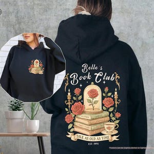 Puede incluir: Sudadera con capucha negra con un dise&ntilde;o floral que incluye una rosa, libros y una taza de t&eacute;. La parte trasera de la sudadera muestra "Belle's Book Club" con un dise&ntilde;o de rosa y vela, y el texto "Tale as Old as Time, Est. 1991".