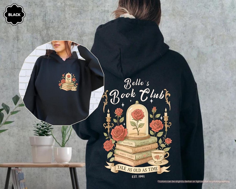 Puede incluir: Sudadera con capucha negra con un dise&ntilde;o floral que incluye una rosa, libros y una taza de t&eacute;. La parte trasera de la sudadera muestra "Belle's Book Club" con un dise&ntilde;o de rosa y vela, y el texto "Tale as Old as Time, Est. 1991".