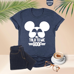 May include: A heather navy blue t-shirt with a white Mickey Mouse design wearing sunglasses and the text "TALK TO ME GOOF". The image also includes a pair of dark blue jeans, a coffee cup, and glasses.