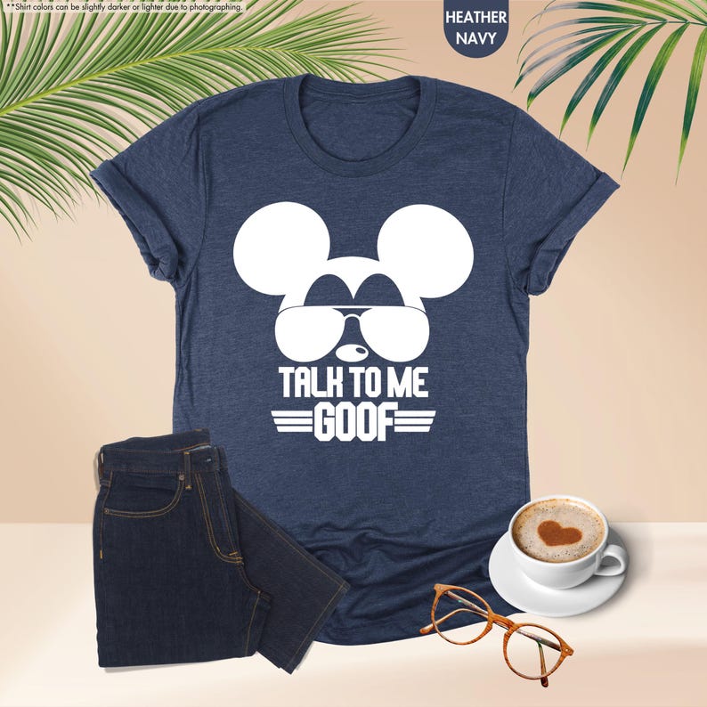 May include: A heather navy blue t-shirt with a white Mickey Mouse design wearing sunglasses and the text "TALK TO ME GOOF". The image also includes a pair of dark blue jeans, a coffee cup, and glasses.