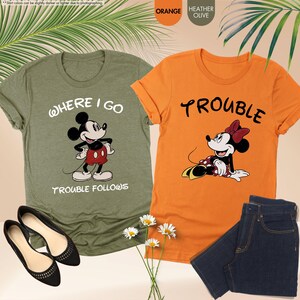 May include: Two graphic t-shirts featuring Disney characters. One is olive green with Mickey Mouse and the text "WHERE I GO TROUBLE FOLLOWS." The other is orange with Minnie Mouse and the word "TROUBLE." Also shown are a pair of black flats and a pair of blue jeans.