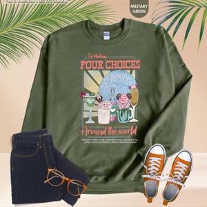 Pu&ograve; includere: Felpa verde militare con la scritta "I'm Making Pour Choices Around the World" e una grafica di cocktail e una sfera blu. La felpa &egrave; abbinata a jeans blu scuro e scarpe da ginnastica arancioni.