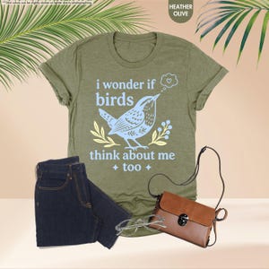 May include: A heather olive t-shirt featuring a light blue graphic of a bird with the text "i wonder if birds think about me too" and a small heart in a thought bubble, accompanied by dark blue jeans, a brown crossbody bag, and clear glasses.