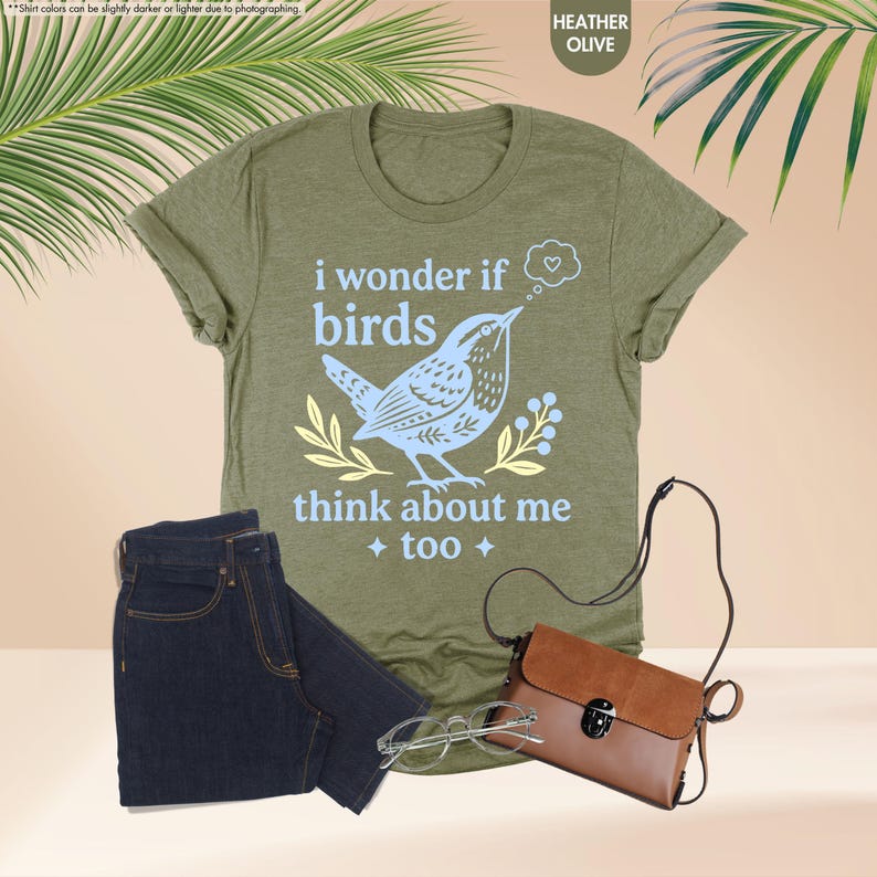 May include: A heather olive t-shirt featuring a light blue graphic of a bird with the text "i wonder if birds think about me too" and a small heart in a thought bubble, accompanied by dark blue jeans, a brown crossbody bag, and clear glasses.