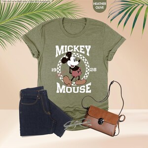 May include: Heather olive green t-shirt featuring a graphic of Mickey Mouse with the text "MICKEY MOUSE 1928". The image also includes a pair of dark blue jeans, a brown purse, and eyeglasses. The shirt is folded.