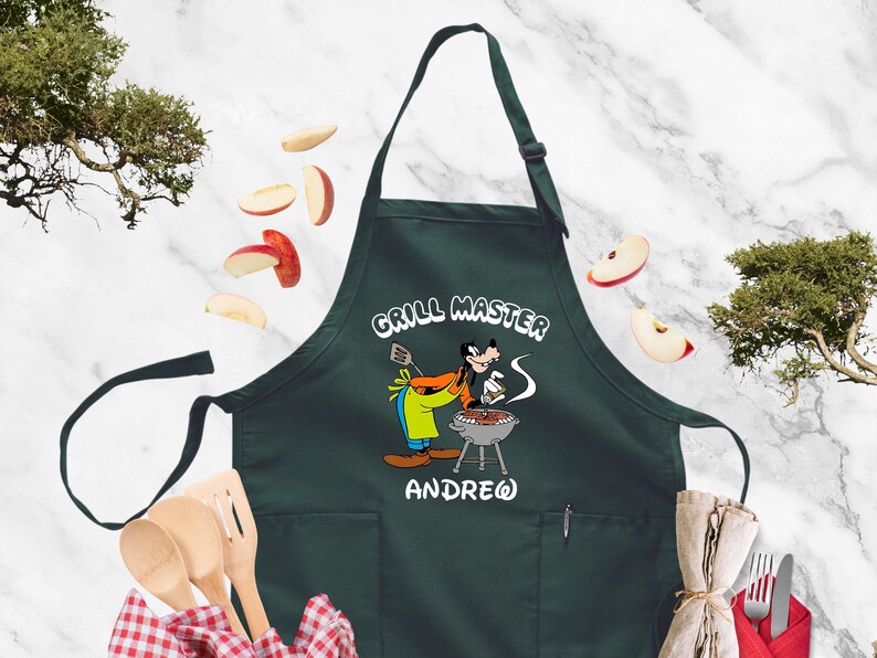 May include: Dark green apron with the words "GRILL MASTER" and a cartoon character grilling. The apron has a pocket and the name "ANDREW" below the character. Slices of apples are scattered around the apron.