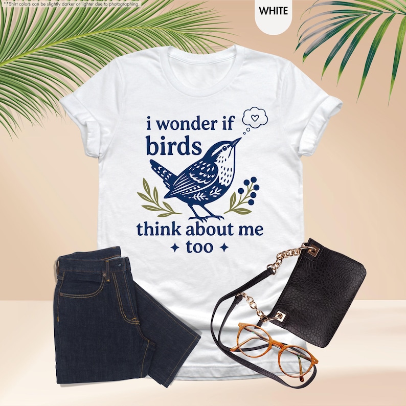 May include: A white short-sleeve t-shirt featuring a dark blue graphic of a bird and the text "i wonder if birds think about me too" with a thought bubble containing a heart, accompanied by dark blue jeans, a black crossbody bag, and glasses.
