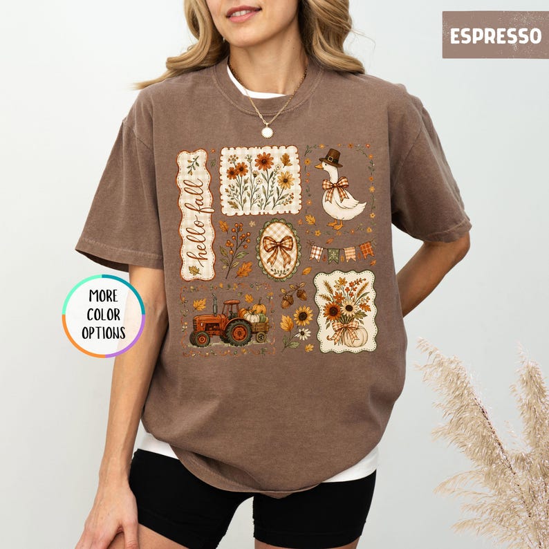May include: An espresso-colored t-shirt featuring a graphic print with the text "hello fall". The design includes a tractor, a duck wearing a hat, floral arrangements, a bow, and autumn leaves. A circular badge on the left reads "MORE COLOR OPTIONS".