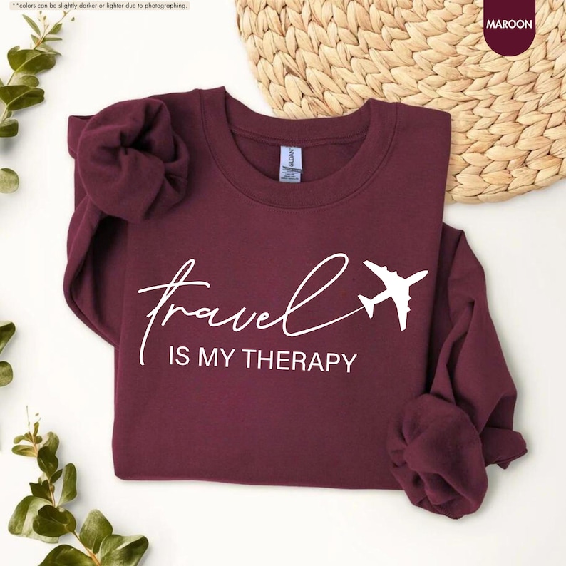 Pu&ograve; includere: Felpa bordeaux con la scritta bianca "travel is my therapy" e una grafica di aereo bianca. Il top girocollo a maniche lunghe &egrave; esposto su una superficie bianca.