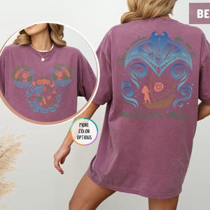 May include: A berry colored t-shirt with a blue and pink graphic design on the back. The graphic features a stylized image of a boat with a person in it. The text "There is nowhere you can go that I won't be with you" is printed on the shirt.