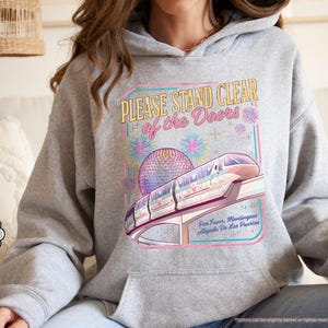 May include: Sport grey hoodie featuring a colorful graphic of a monorail, a geodesic sphere, and fireworks. The text on the graphic reads "PLEASE STAND CLEAR of the Doors" and "Por Favor, Mantingase Alejados De Las Puertas."
