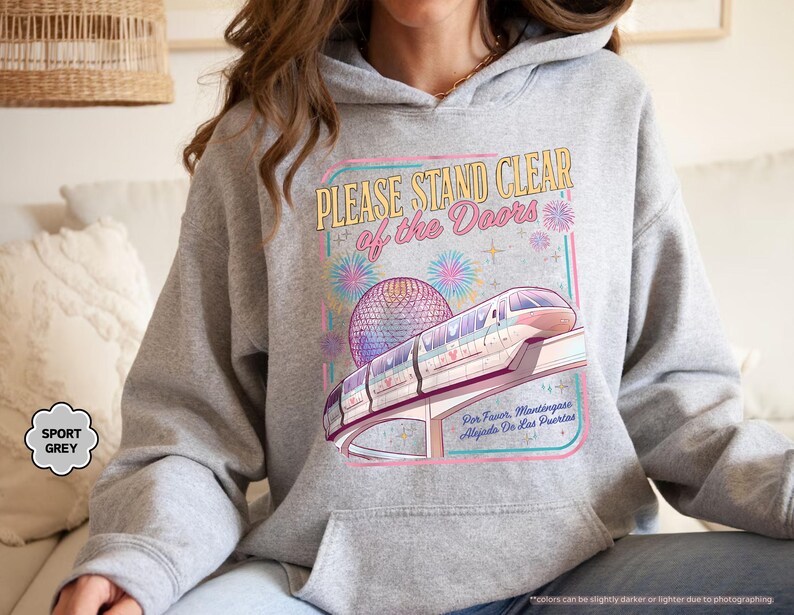 May include: Sport grey hoodie featuring a colorful graphic of a monorail, a geodesic sphere, and fireworks. The text on the graphic reads "PLEASE STAND CLEAR of the Doors" and "Por Favor, Mantingase Alejados De Las Puertas."