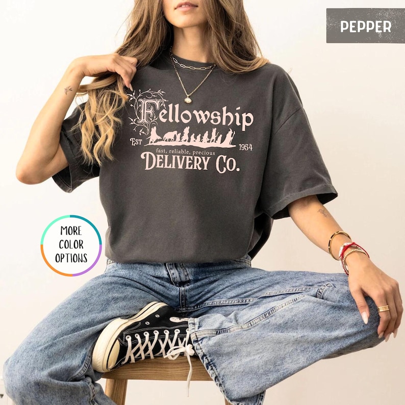 May include: A gray t-shirt with a vintage-style graphic that says "Fellowship Delivery Co." with the text "Est. 1954" and "fast, reliable, precious" underneath. The graphic features a line of small figures walking through a forest.