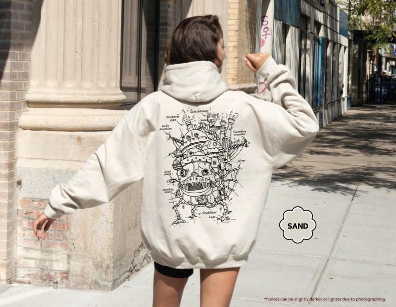 May include: A cream-colored hoodie with a black and white illustration of a flying machine on the back. The illustration includes text labels for different parts of the machine. The text "SAND" is in a circle on the bottom right corner of the image.
