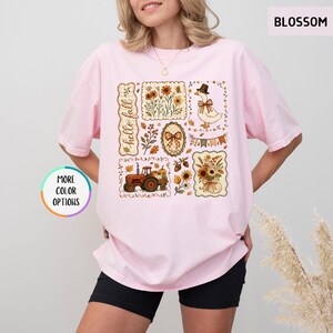 May include: A light pink short-sleeve t-shirt featuring a graphic print with the text "hello fall". The design includes a red tractor, a goose wearing a hat, sunflowers, autumn leaves, pumpkins, and plaid bows arranged in a collage style.
