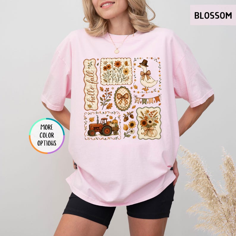 May include: A light pink short-sleeve t-shirt featuring a graphic print with the text "hello fall". The design includes a red tractor, a goose wearing a hat, sunflowers, autumn leaves, pumpkins, and plaid bows arranged in a collage style.