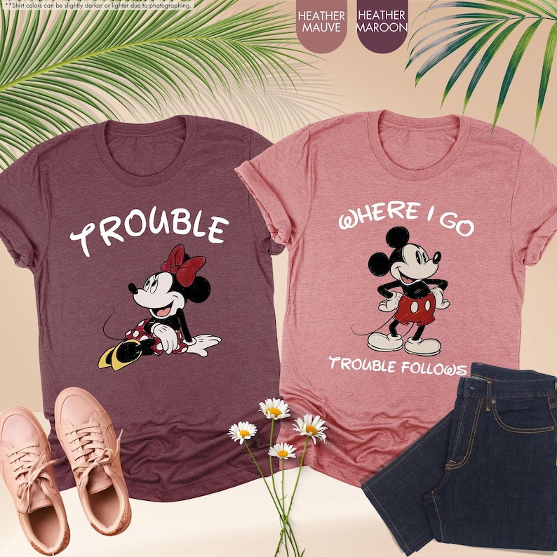 May include: Two graphic t-shirts, one maroon and one mauve, featuring Disney's Minnie and Mickey Mouse. The maroon shirt says "Trouble" with Minnie Mouse. The mauve shirt says "Where I Go Trouble Follows" with Mickey Mouse. Pink sneakers and jeans are also shown.