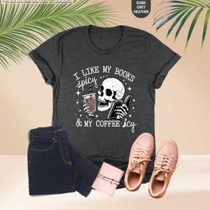 May include: A dark heather grey t-shirt with a white graphic of a skeleton holding a book and a cup of coffee. The text on the shirt reads "I like my books spicy & my coffee icy".