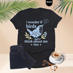 May include: A black short-sleeved t-shirt featuring a light blue graphic of a bird with the text "i wonder if birds think about me too" and a thought bubble containing a heart, accompanied by yellow leaf accents and four-pointed stars.