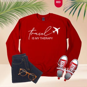 Pu&ograve; includere: Una felpa rossa con la scritta "travel is my therapy" e un'immagine di un aereo bianco. L'immagine mostra anche un paio di jeans blu, occhiali con montatura dorata e scarpe da ginnastica rosse.