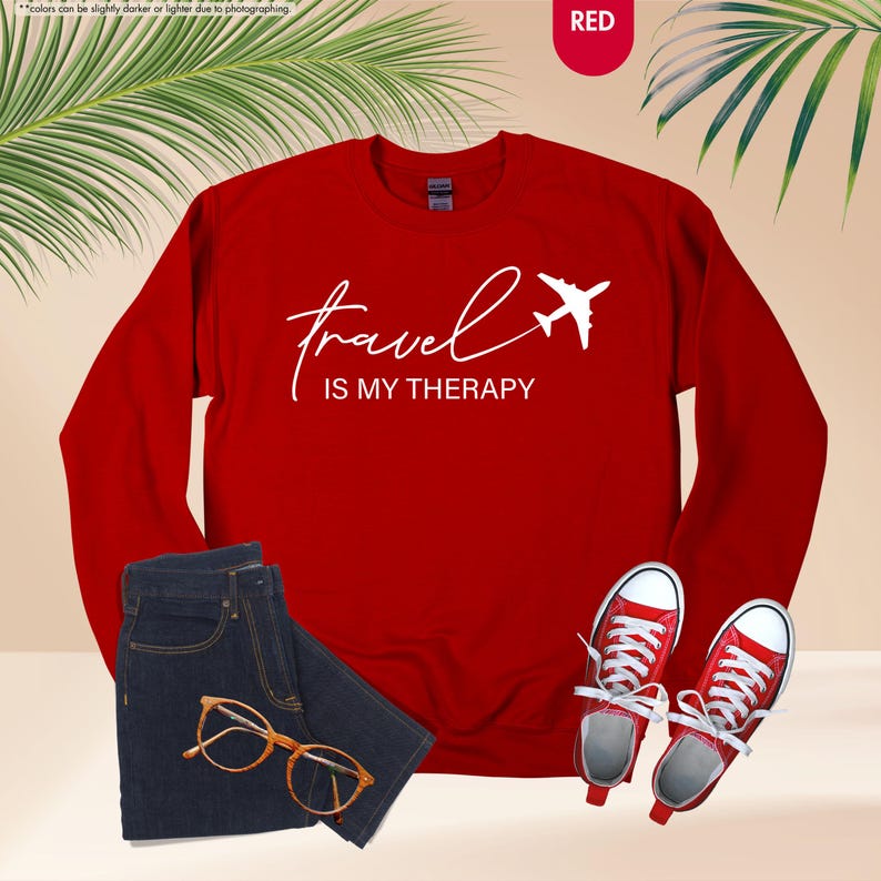 Pu&ograve; includere: Una felpa rossa con la scritta "travel is my therapy" e un'immagine di un aereo bianco. L'immagine mostra anche un paio di jeans blu, occhiali con montatura dorata e scarpe da ginnastica rosse.