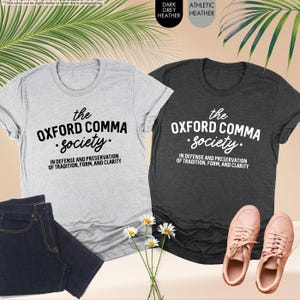 Op de afbeelding: Twee T-shirts met de tekst "the Oxford comma society. In defense and preservation of tradition, form, and clarity" erop gedrukt. E&eacute;n shirt is donkergrijs gem&ecirc;leerd en de andere is lichtgrijs gem&ecirc;leerd.