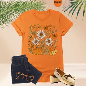 May include: Orange t-shirt featuring a floral and pumpkin design. The shirt is styled with dark blue jeans, tan loafers, and orange-framed glasses. The design includes daisies, pumpkins, and other autumn-themed elements.