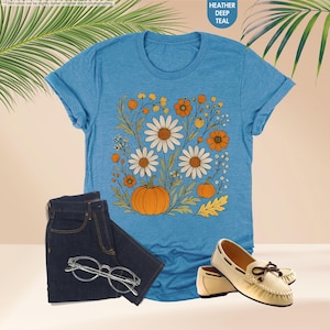 May include: A teal t-shirt with a floral and pumpkin design, alongside dark wash jeans, eyeglasses, and cream-coloured loafers. The shirt features white daisies, orange flowers, and pumpkins. The text "HEATHER DEEP TEAL" is in the upper right corner.