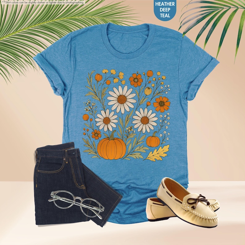 May include: A teal t-shirt with a floral and pumpkin design, alongside dark wash jeans, eyeglasses, and cream-coloured loafers. The shirt features white daisies, orange flowers, and pumpkins. The text "HEATHER DEEP TEAL" is in the upper right corner.