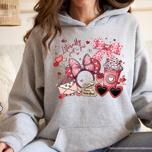 May include: A light grey hoodie featuring a Valentine's Day design. The design includes a pink bow, heart-shaped sunglasses, a coffee cup, and the text "Literally just a girl." The hoodie is labeled "Sport Grey."