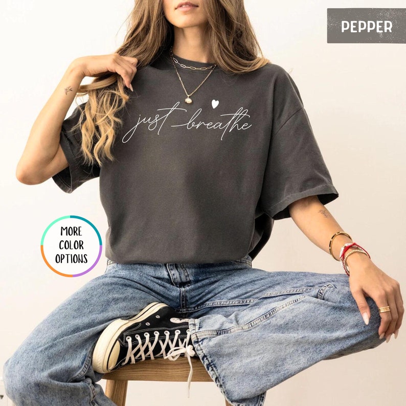 May include: A gray t-shirt with the text "just breathe" in white script with a small white heart above the text. The model is wearing the shirt with blue jeans and black sneakers.