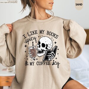 May include: A beige sweatshirt with a black and white graphic print of a skull holding a book and a cup of coffee. The text on the sweatshirt reads "I like my books spicy & my coffee icy".