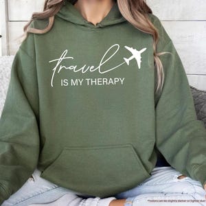 Pu&ograve; includere: Una felpa con cappuccio verde oliva con la scritta bianca "travel is my therapy" e un'immagine di un aereo bianco. La felpa ha una tasca frontale e un cappuccio. Le parole "MILITARY GREEN" sono in una piccola grafica a sinistra.