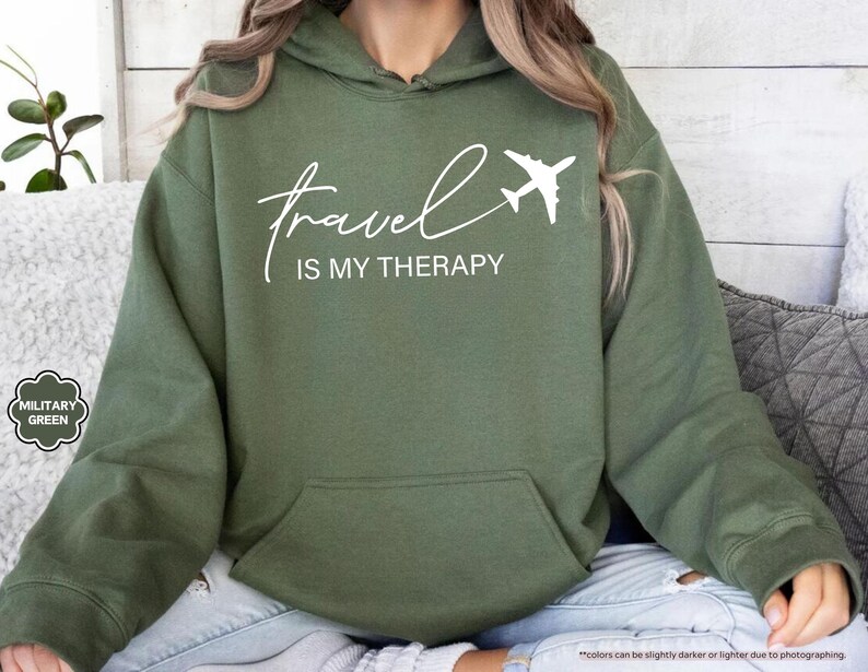 Pu&ograve; includere: Una felpa con cappuccio verde oliva con la scritta bianca "travel is my therapy" e un'immagine di un aereo bianco. La felpa ha una tasca frontale e un cappuccio. Le parole "MILITARY GREEN" sono in una piccola grafica a sinistra.