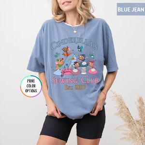 May include: A light blue t-shirt with a graphic of Cinderella's mice sewing. The text reads "Cinderella's Sewing Club Est. 1950".