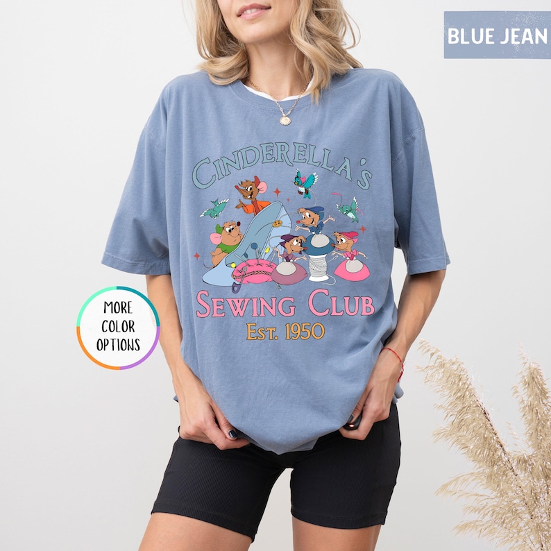 May include: A light blue t-shirt with a graphic of Cinderella's mice sewing. The text reads "Cinderella's Sewing Club Est. 1950".