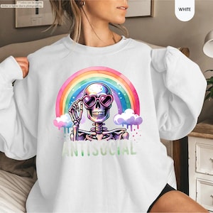 May include: White sweatshirt with a skeleton wearing heart-shaped sunglasses, set against a rainbow backdrop. The word "ANTISOCIAL" is printed in green below the skeleton. The design is vibrant and colourful.
