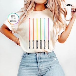 K&ouml;nnte beinhalten: Cremefarbenes T-Shirt mit einem Design aus sechs farbigen Lichtschwertern in Rot, Orange, Gelb, Gr&uuml;n, Blau und Lila. Das Shirt hat den Text "MORE COLOR OPTIONS" in einem Kreis.