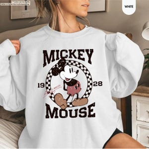 May include: White sweatshirt featuring a vintage Mickey Mouse graphic. The design includes the words "MICKEY MOUSE" and the year "1928" in a retro font. The cartoon character is in the center, with a checkered circle behind it.
