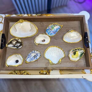 May include: A wooden tray with nine decorative seashells. The shells are painted with various patterns and designs, including floral, geometric, and abstract motifs. The shells are arranged in a circular pattern on the tray, with a gold rim around each shell.